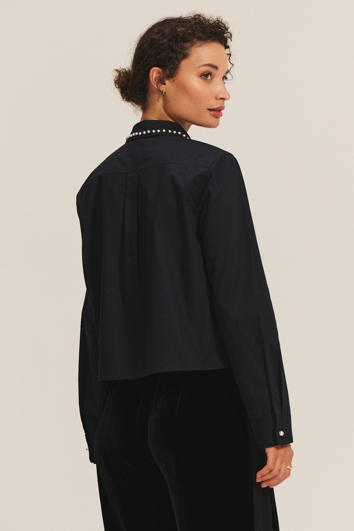 A woman with curly hair wears the JOCELYN SHIRT by Velvet by Graham & Spencer, featuring a black beaded collar with pearl accents, as she stands facing away and looks over her shoulder against a plain background.