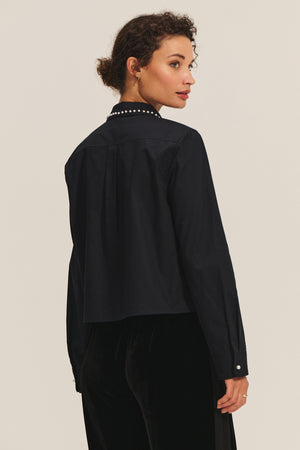 A woman with curly hair wears the JOCELYN SHIRT by Velvet by Graham & Spencer, featuring a black beaded collar with pearl accents, as she stands facing away and looks over her shoulder against a plain background.