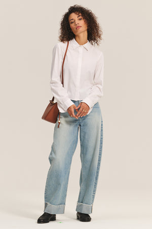 A person stands against a plain background wearing the JOCELYN SHIRT by Velvet by Graham & Spencer, paired with light blue wide-leg jeans with cuffs, black shoes, and carrying a brown shoulder bag.
