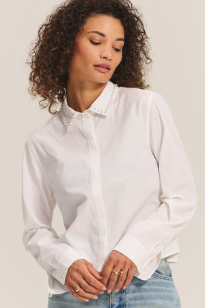 A woman wears the JOCELYN SHIRT by Velvet by Graham & Spencer, a white cotton poplin shirt with a beaded collar, paired with light blue jeans against a plain background.