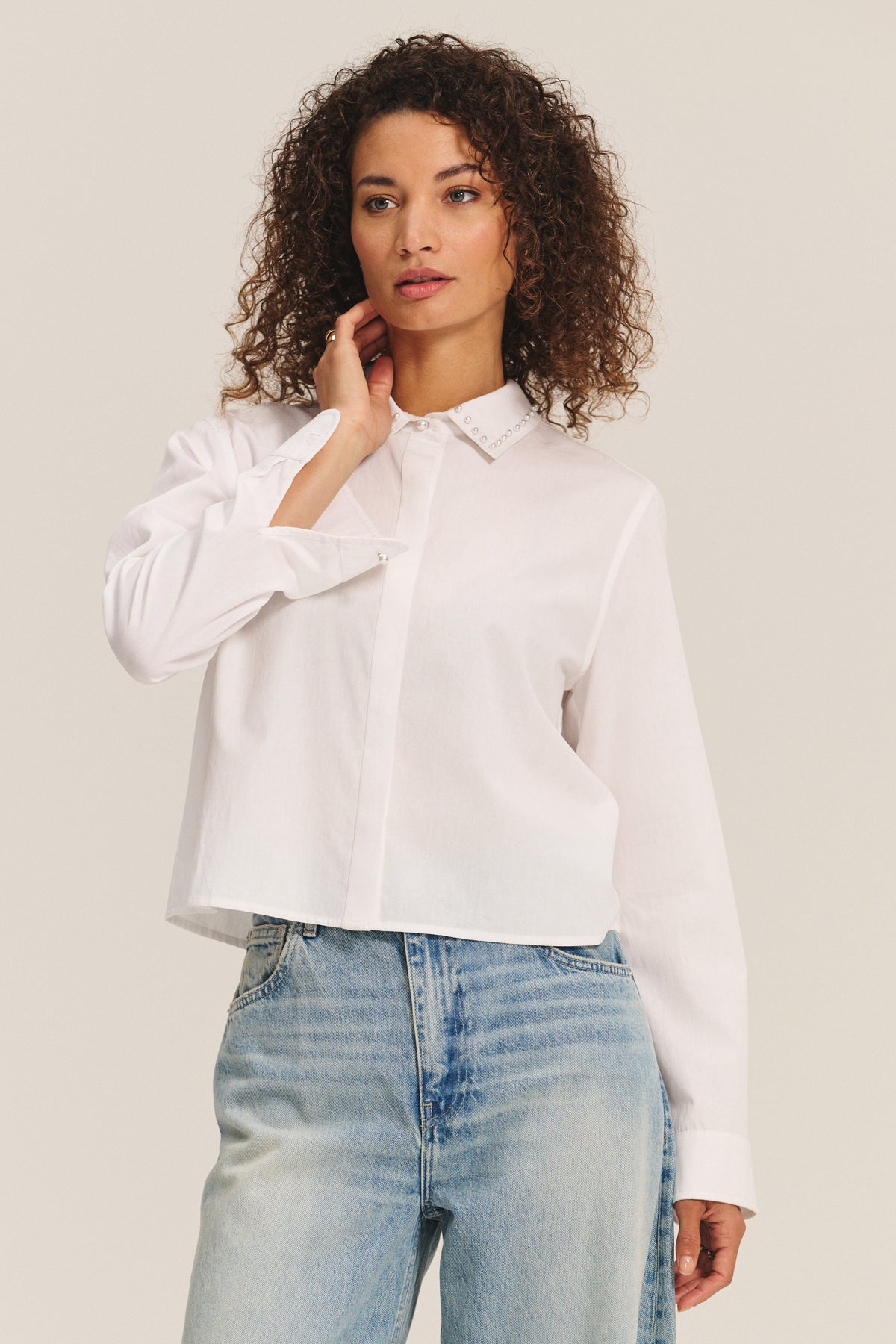A woman with curly hair wears the Velvet by Graham & Spencer JOCELYN SHIRT in white cotton poplin and light blue jeans, standing against a plain background.-40247705501889