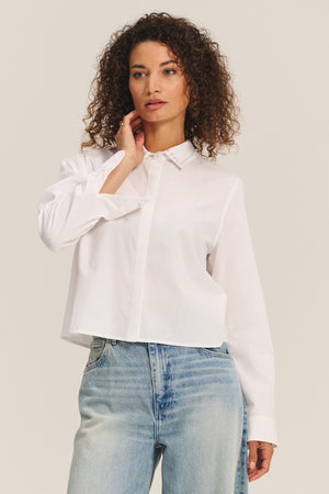 A woman with curly hair wears the Velvet by Graham & Spencer JOCELYN SHIRT in white cotton poplin and light blue jeans, standing against a plain background.