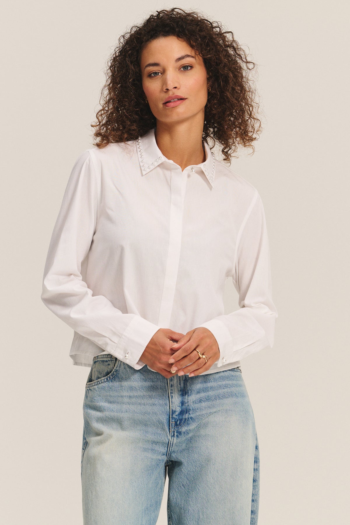 A woman wears the JOCELYN SHIRT by Velvet by Graham & Spencer, a white cotton poplin top with an embroidered collar, paired with light blue jeans and standing against a plain background.-40247705469121