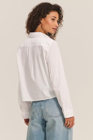 A woman with curly hair wears the JOCELYN SHIRT by Velvet by Graham & Spencer, a white cotton poplin top, paired with light blue jeans as she stands looking over her shoulder.