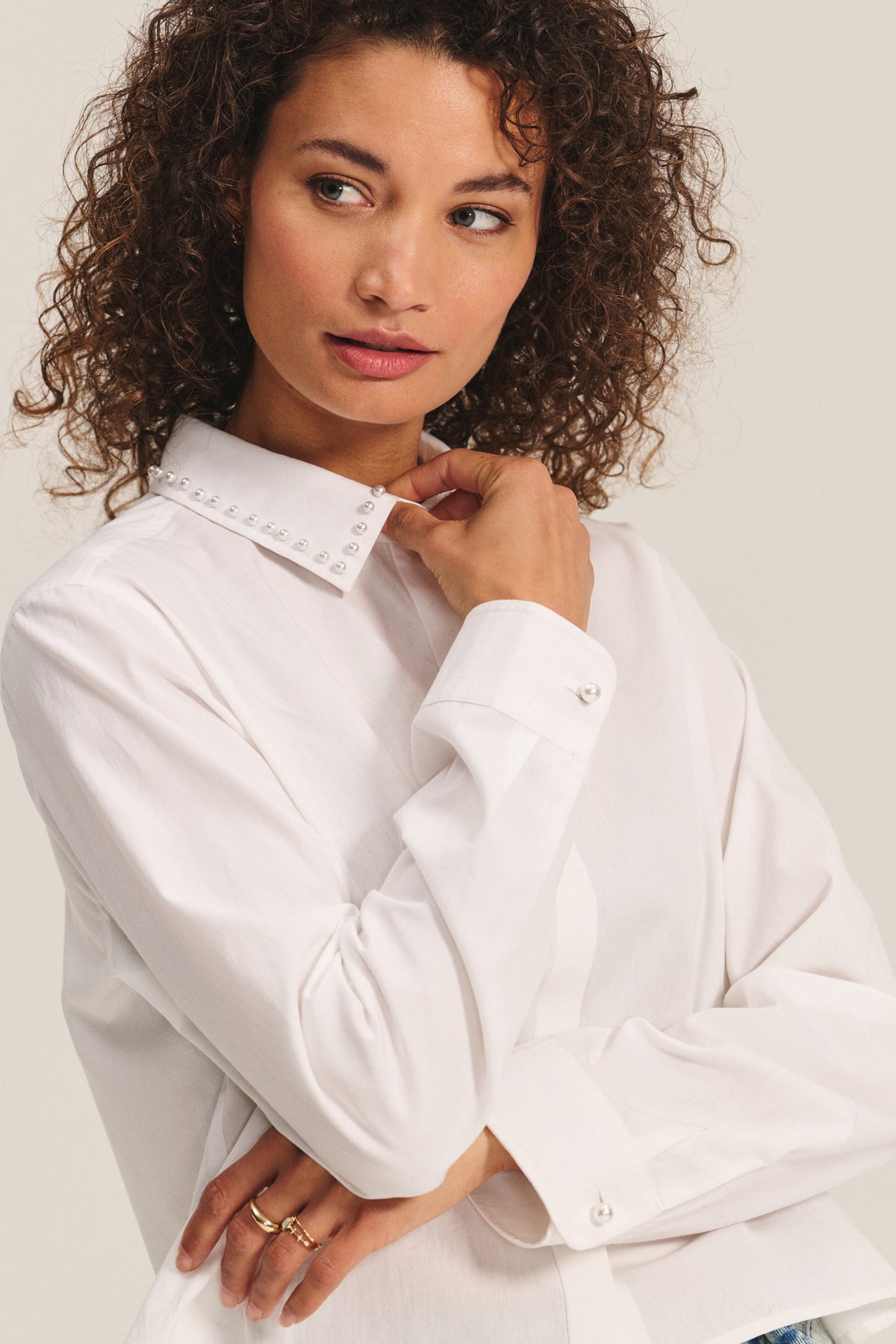 Woman with curly hair wearing the JOCELYN SHIRT by Velvet by Graham & Spencer—a white cotton poplin shirt featuring pearl-embellished collar—looks to the side, one hand gently touching her collar.-40247705403585