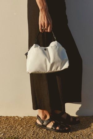 A person wearing a black outfit and black sandals holds a small white NYLON CROSSBODY BAG by Velvet by Graham & Spencer with a secure zipper closure by their side, standing against a plain wall.