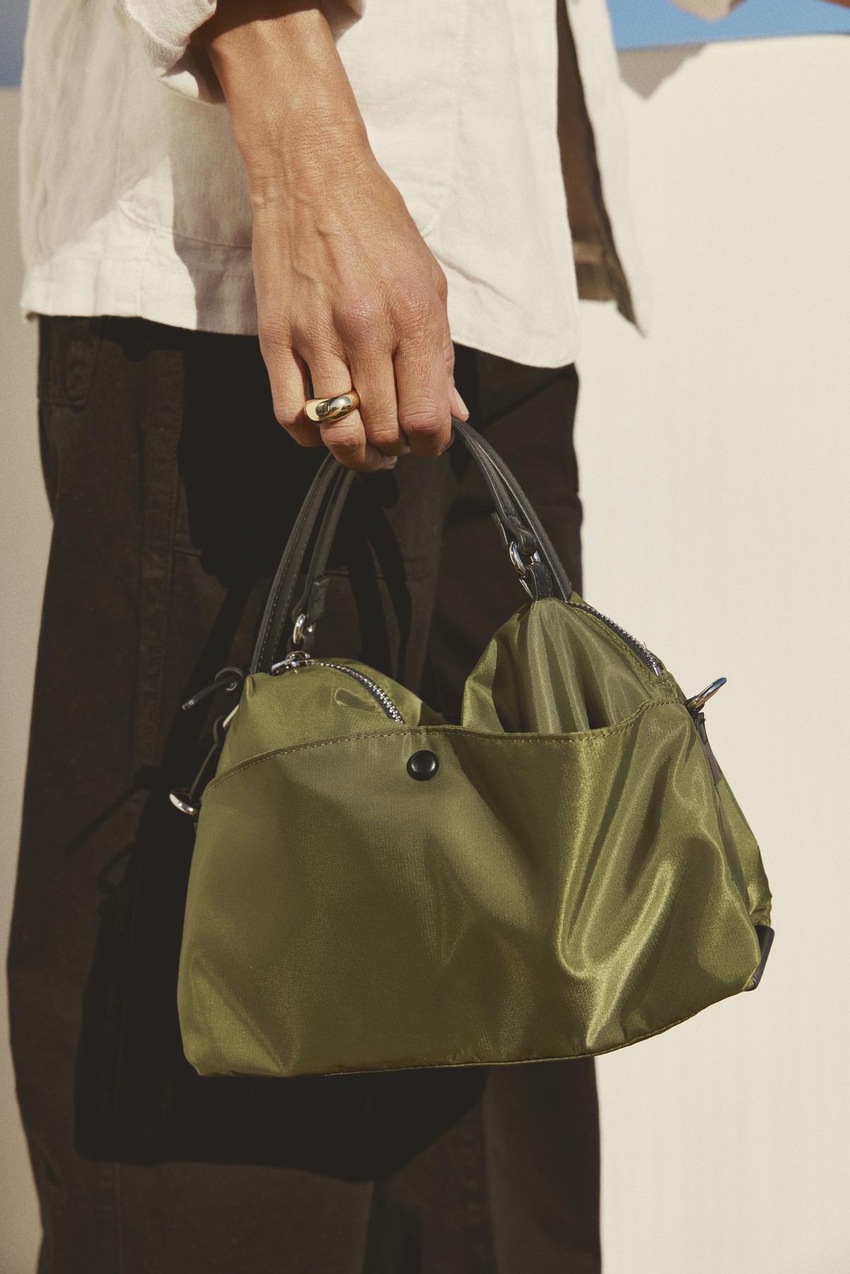   A person holds a small, green handbag with black handles. Their fingers are adorned with a gold ring. They are dressed in a light-colored shirt and dark pants. The Velvet by Graham & Spencer NYLON CROSSBODY BAG features a secure zipper closure, crafted from durable nylon for everyday reliability.
 