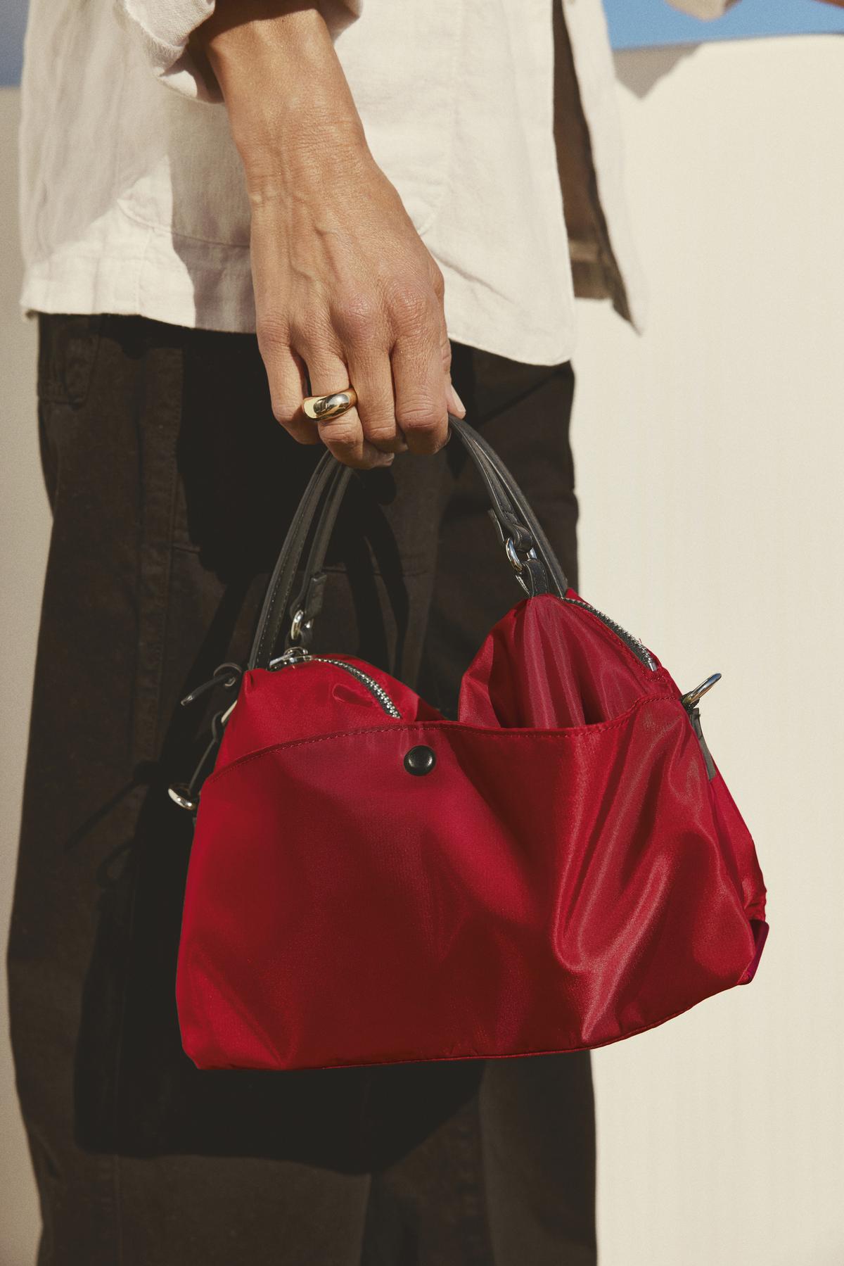   A person carries the NYLON CROSSBODY BAG from Velvet by Graham & Spencer, featuring a compact design in a vibrant red nylon with black handles, paired elegantly with a silver ring, black trousers, and a light-colored top—perfect for daily wear. 