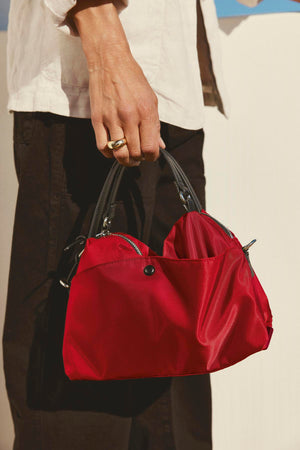 A person carries the NYLON CROSSBODY BAG from Velvet by Graham & Spencer, featuring a compact design in a vibrant red nylon with black handles, paired elegantly with a silver ring, black trousers, and a light-colored top—perfect for daily wear.