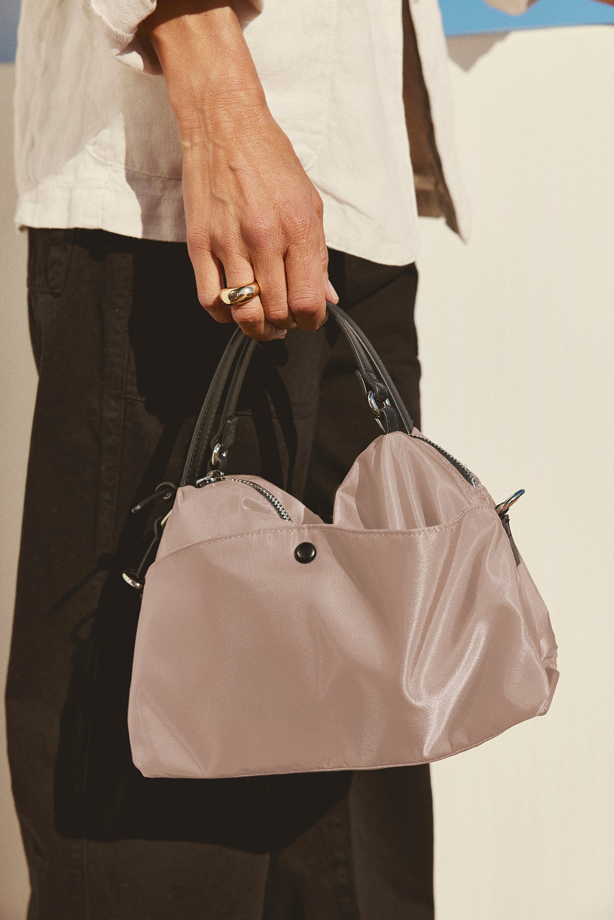   A person carries a Velvet by Graham & Spencer NYLON CROSSBODY BAG in light pink with black handles, crafted from durable nylon, ideal for everyday use. They're dressed in a white shirt and black pants. 