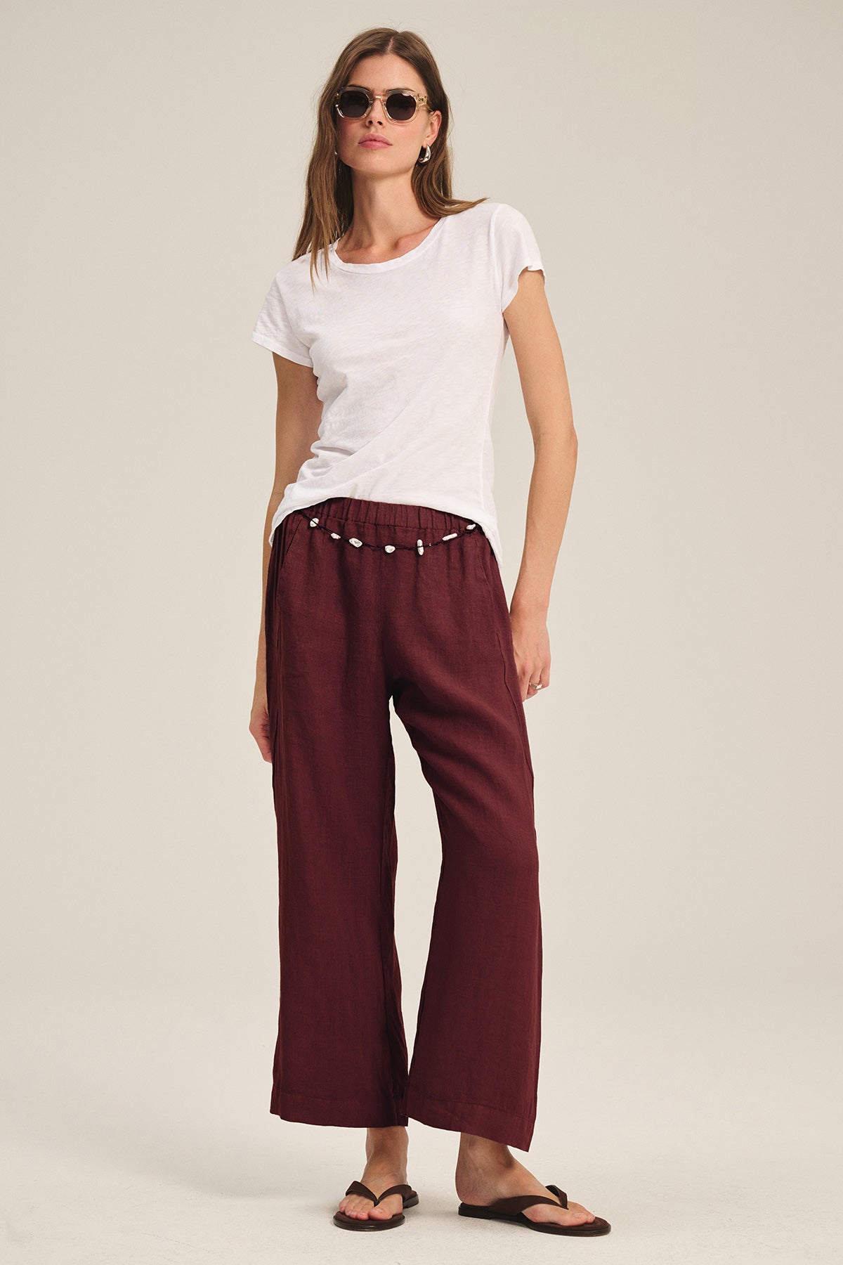 A woman stands against a plain background wearing sunglasses, a white t-shirt, black sandals, and Velvet by Graham & Spencer's LOLA LINEN PANT in maroon with an elastic waist.