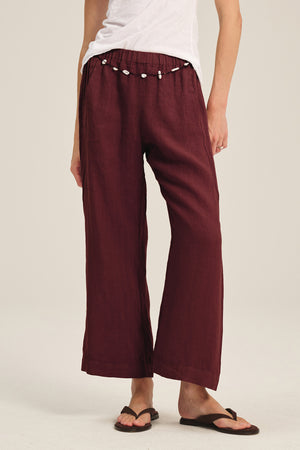 Person wearing white t-shirt, Velvet by Graham & Spencer LOLA LINEN PANT in burgundy with elastic waist and shell belt, paired with black flip-flops, against a neutral background.