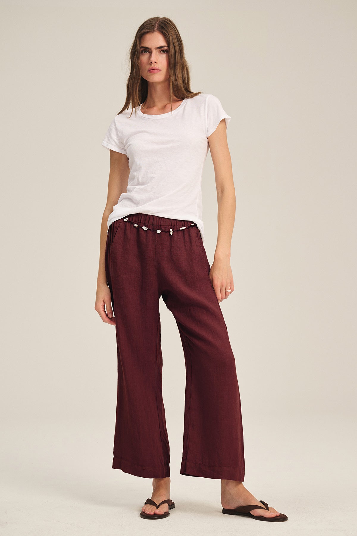A woman stands against a plain background wearing a white t-shirt, Velvet by Graham & Spencer's LOLA LINEN PANT in maroon with an elastic waist, and black flip-flops.-40009910452417