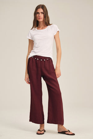 A woman stands against a plain background wearing a white t-shirt, Velvet by Graham & Spencer's LOLA LINEN PANT in maroon with an elastic waist, and black flip-flops.