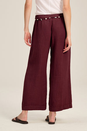 A person is shown from the back wearing the LOLA LINEN PANT by Velvet by Graham & Spencer—wide-leg maroon linen trousers featuring a shell-embellished elastic waist—paired with a white shirt and black sandals.