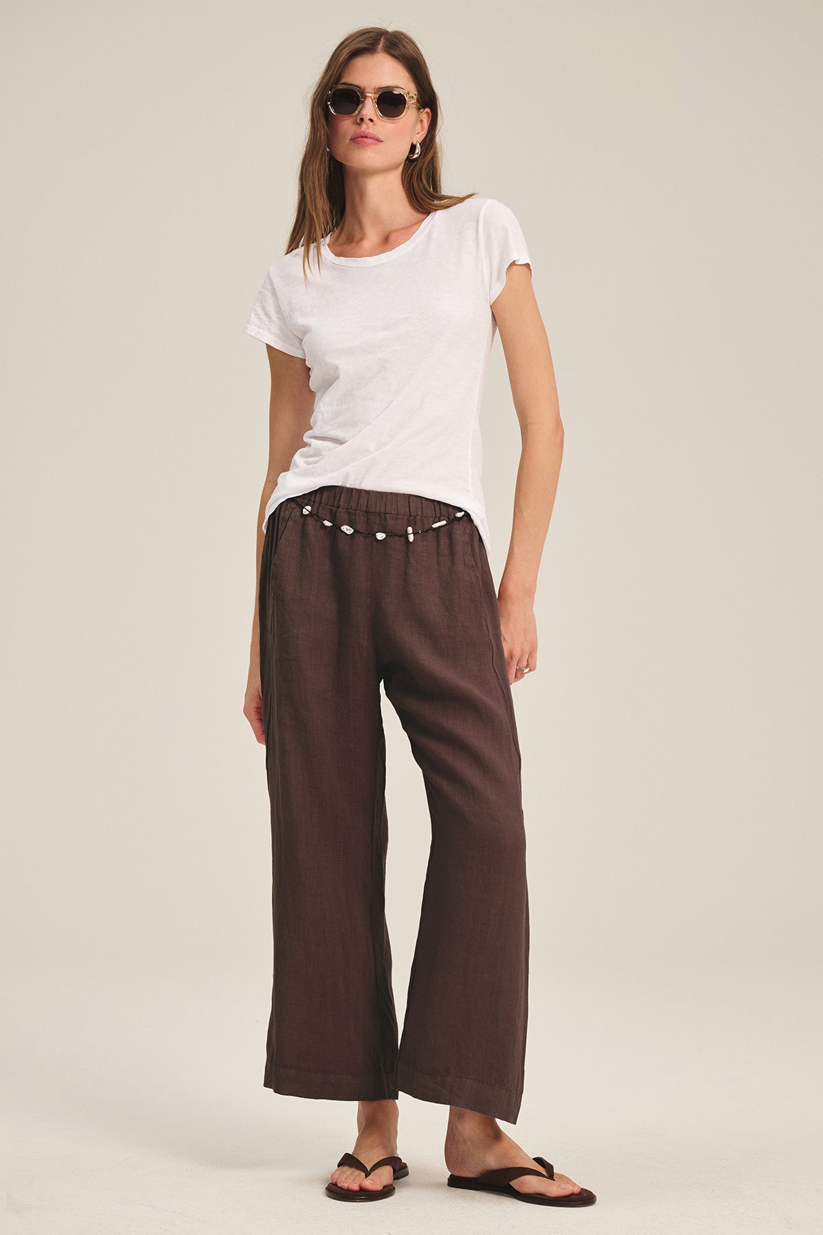 Woman wearing sunglasses, a white t-shirt, Velvet by Graham & Spencer LOLA LINEN PANT in brown with an elastic waist and drawstring, and black flip-flops, standing against a plain background.-40009910485185