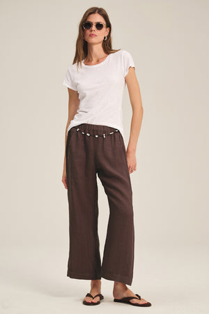 Woman wearing sunglasses, a white t-shirt, Velvet by Graham & Spencer LOLA LINEN PANT in brown with an elastic waist and drawstring, and black flip-flops, standing against a plain background.