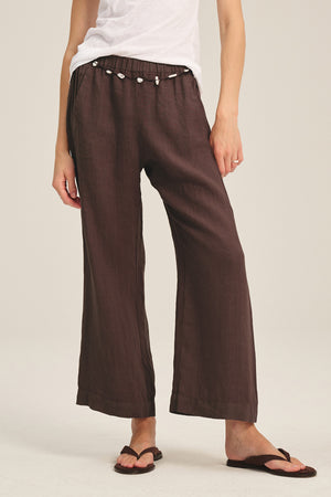 A person stands against a plain background wearing Velvet by Graham & Spencer's LOLA LINEN PANT—lightweight linen trousers with an elastic waist and shell belt—paired with a white T-shirt and black flip-flops.