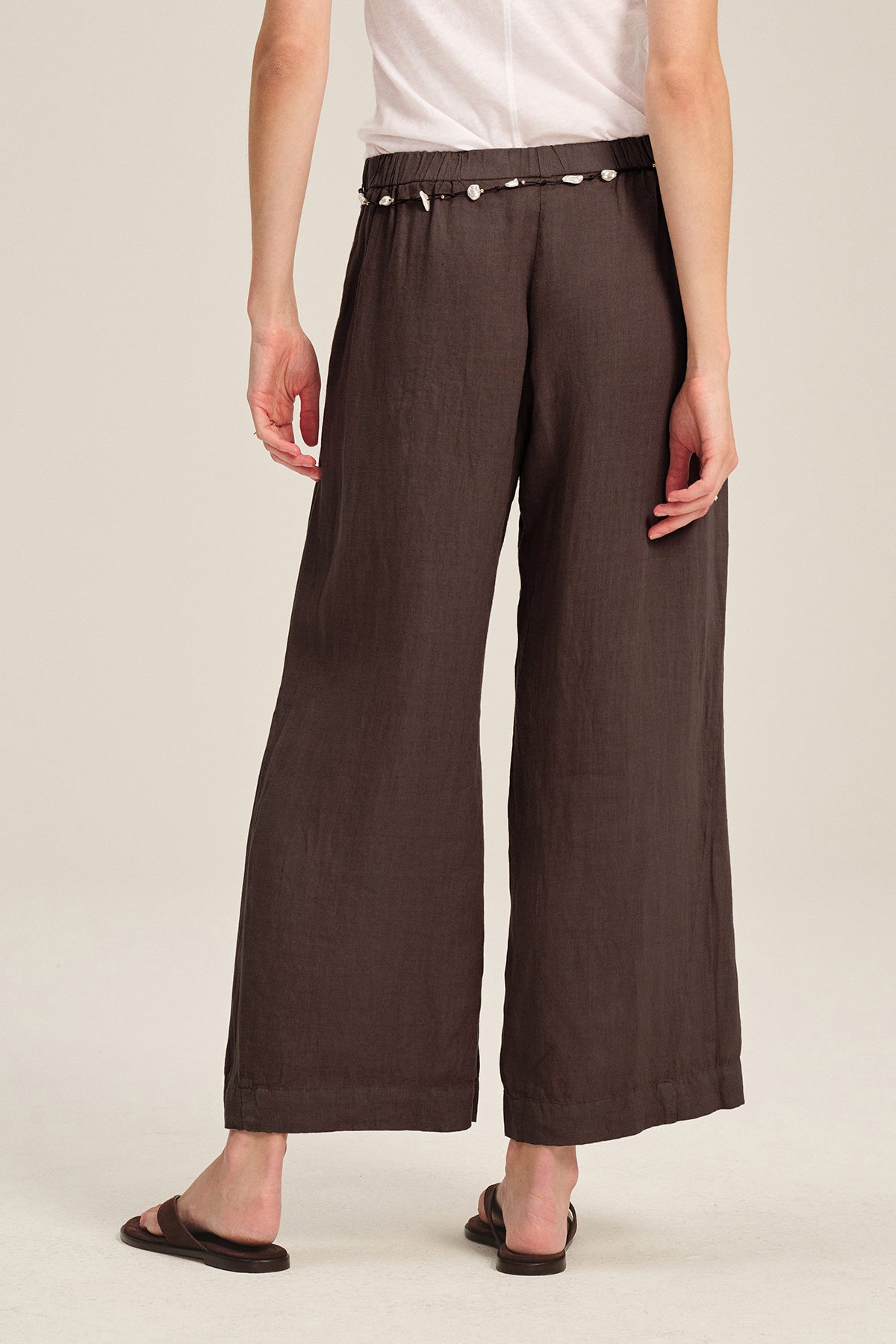 A person wearing the LOLA LINEN PANT by Velvet by Graham & Spencer—brown, wide-leg, pull-on pants with an elastic waist—paired with a white short-sleeve shirt, black sandals, and a shell belt stands against a plain background.-40012876120257