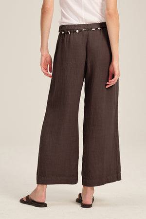 A person wearing the LOLA LINEN PANT by Velvet by Graham & Spencer—brown, wide-leg, pull-on pants with an elastic waist—paired with a white short-sleeve shirt, black sandals, and a shell belt stands against a plain background.