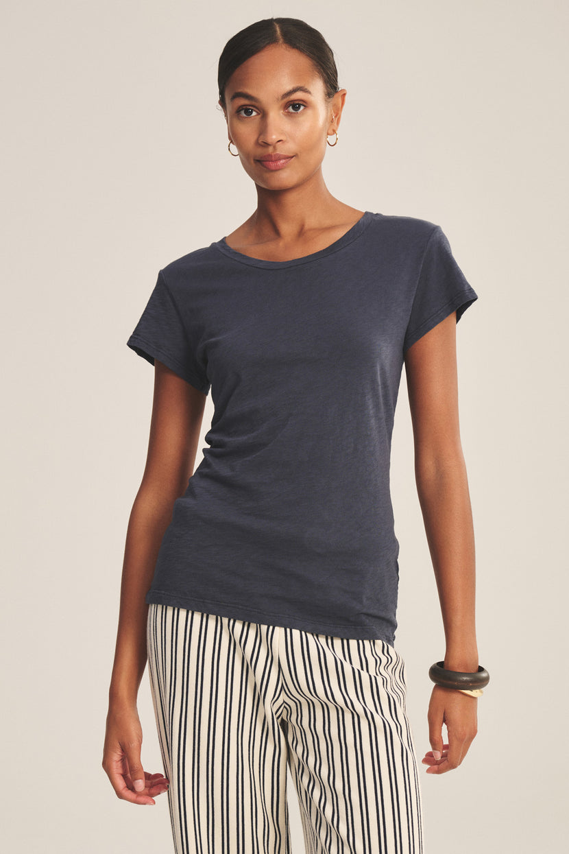 A person stands facing forward, wearing the ODELIA TEE by Velvet by Graham & Spencer in navy cotton slub with a crew neckline, paired with white pants featuring black vertical stripes and accessorized with a black bracelet and small hoop earrings.