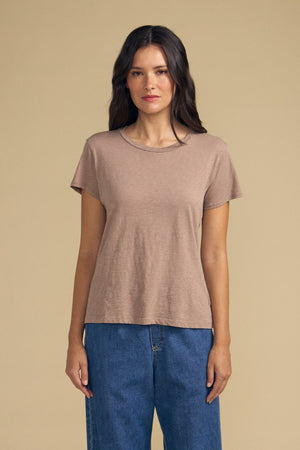 A woman with long brown hair wears the Velvet by Graham & Spencer ODELIA TEE, a plain tan short-sleeve cotton slub top with a classic crew neckline, paired with blue jeans and standing against a beige background.