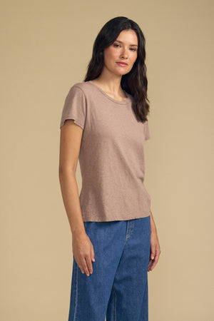 A woman stands against a beige background wearing the ODELIA TEE by Velvet by Graham & Spencer—a light brown premium cotton top featuring a classic crew neckline—paired with blue jeans, looking at the camera with a neutral expression for an effortlessly stylish wardrobe essential.