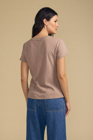 A woman stands facing away, wearing the ODELIA TEE by Velvet by Graham & Spencer—a premium cotton top featuring a classic crew neckline—in taupe. Paired with blue denim jeans, she poses against a plain beige background. This short-sleeve tee is the perfect wardrobe essential.
