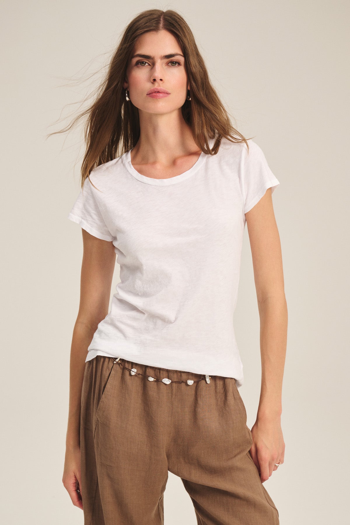 A woman wears the ODELIA TEE by Velvet by Graham & Spencer—a classic crew neck cotton slub tee in plain white—paired with light brown pants, standing against a neutral background. A true wardrobe essential.-40305622679745