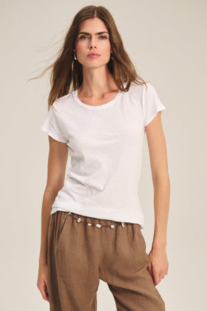 A woman wears the ODELIA TEE by Velvet by Graham & Spencer—a classic crew neck cotton slub tee in plain white—paired with light brown pants, standing against a neutral background. A true wardrobe essential.