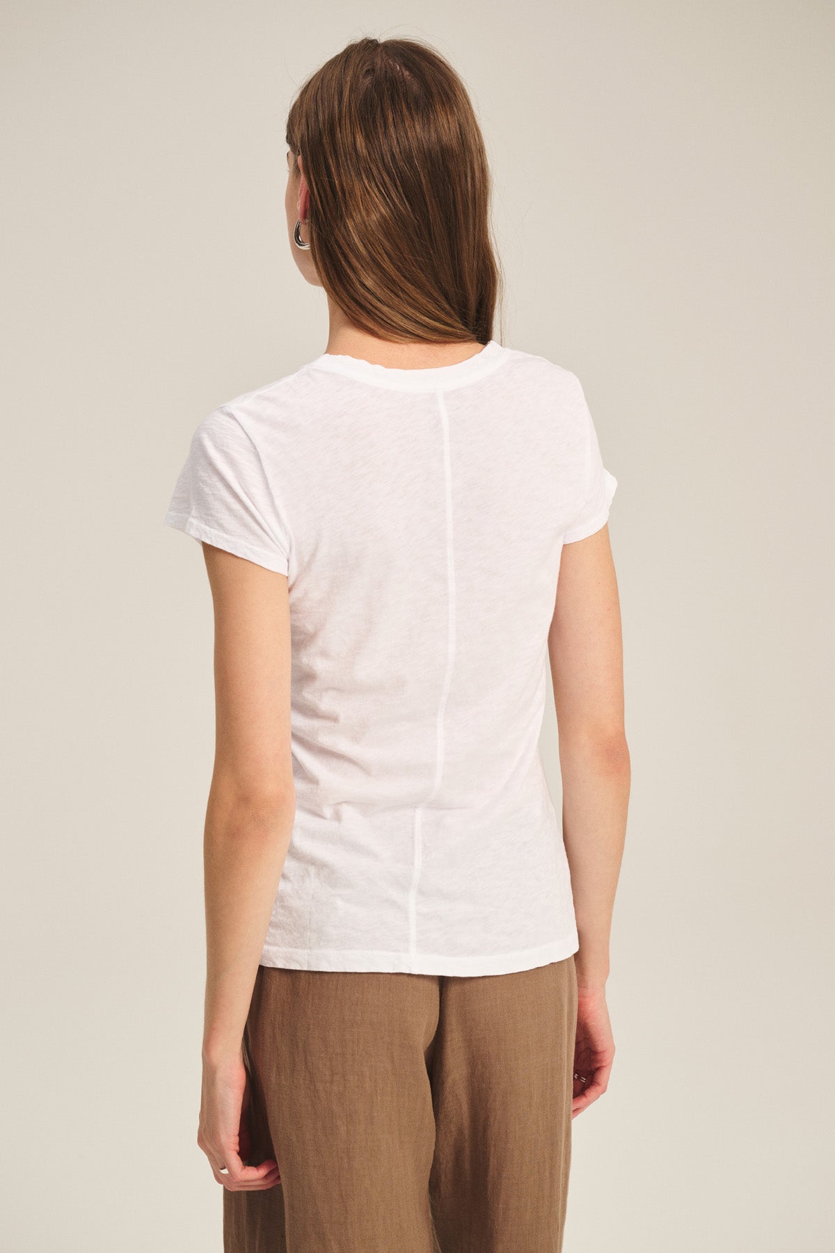 A person with long brown hair, wearing the ODELIA TEE by Velvet by Graham & Spencer and brown pants, stands facing away from the camera against a plain background—a true wardrobe essential.
