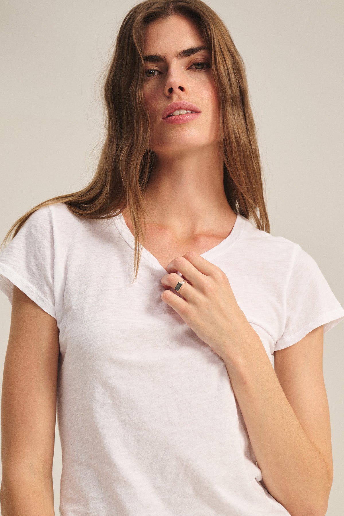 A woman with long brown hair wears the Velvet by Graham & Spencer ODELIA TEE, a classic crew neck cotton slub style, looking at the camera and holding a ring near her chest.