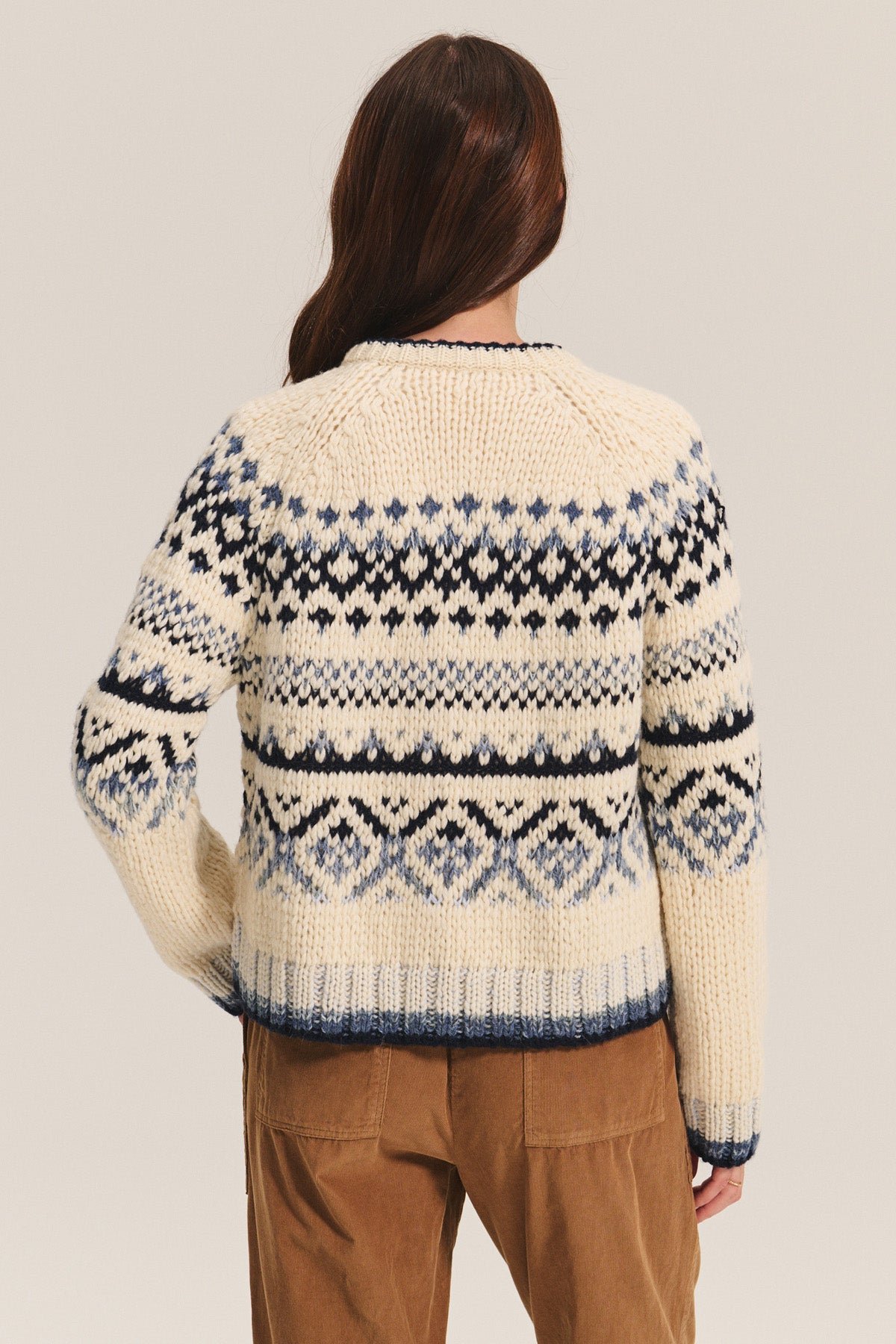 A person faces away, wearing the Velvet by Graham & Spencer TOVAH CARDIGAN—a cream Fair Isle sweater with blue and black geometric patterns—paired with brown corduroy pants against a plain background.-40333274054849