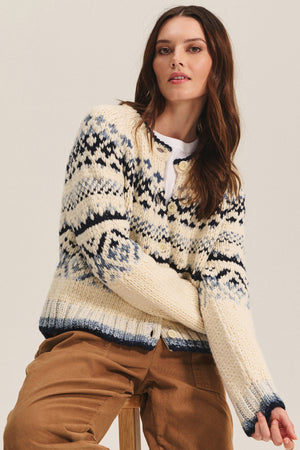 A woman in the Velvet by Graham & Spencer TOVAH CARDIGAN, a cream and navy chunky knit Fair Isle sweater, paired with a white shirt and tan pants, sits on a wooden stool against a plain background.