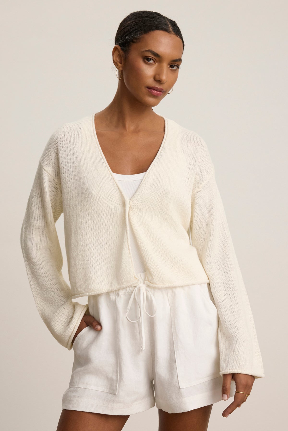 A woman wears the VERITY COTTON CASHMERE MESH CARDIGAN by Velvet by Graham & Spencer over a white tank and drawstring shorts—an ideal transitional look—standing against a neutral background.
