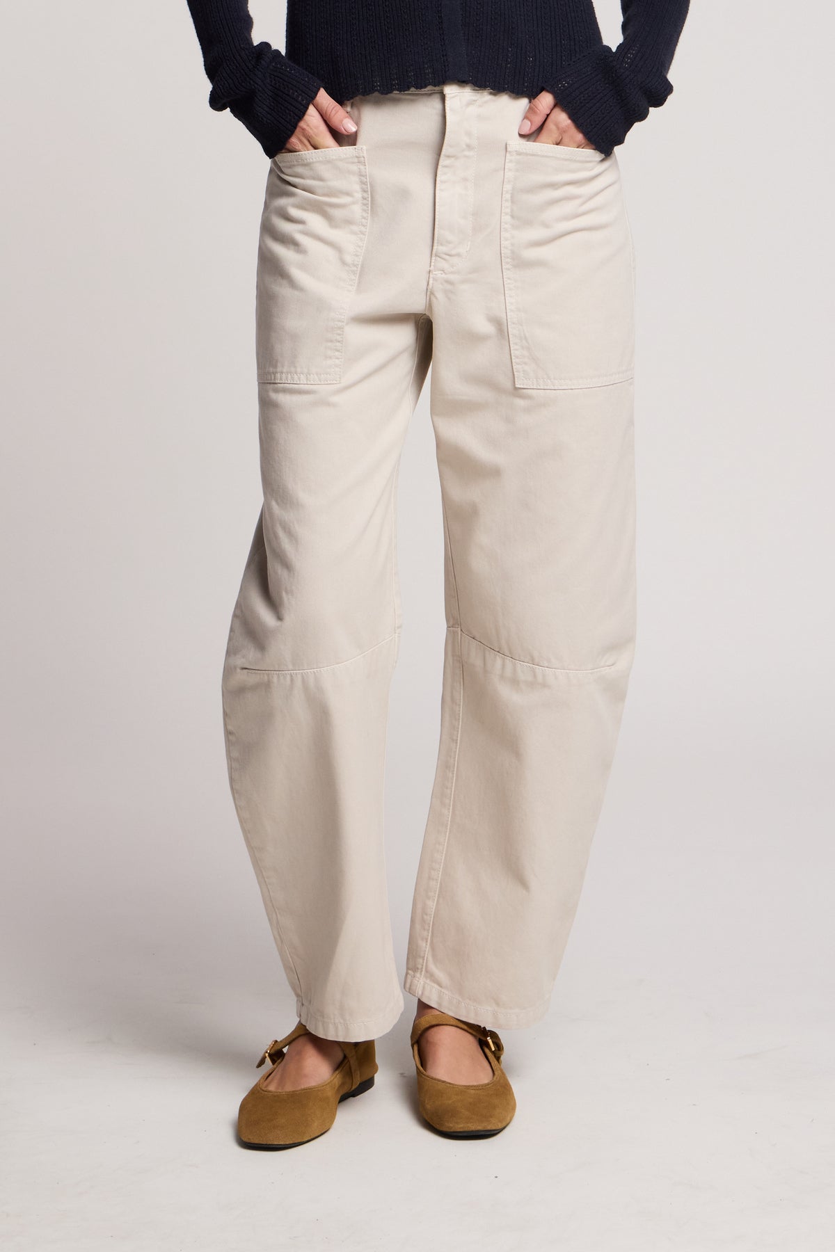 Someone stands against a plain background wearing the BRYLIE PANT by Velvet by Graham & Spencer—light beige cotton twill barrel-leg pants with utilitarian patch pockets—paired with a dark sweater and brown shoes.