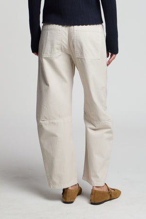 A person stands with their back to the camera, wearing Velvet by Graham & Spencer’s BRYLIE PANT in off-white with patch pockets, a dark long-sleeve top, and brown lace-up shoes against a plain background.