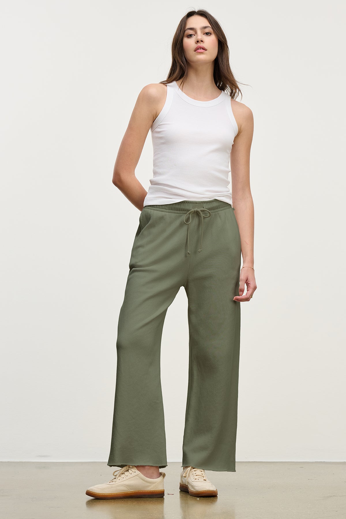   A person is wearing a white sleeveless top, paired with the MONTECITO SWEATPANT by Velvet by Jenny Graham in olive green organic cotton, and beige shoes, standing against a plain background. 