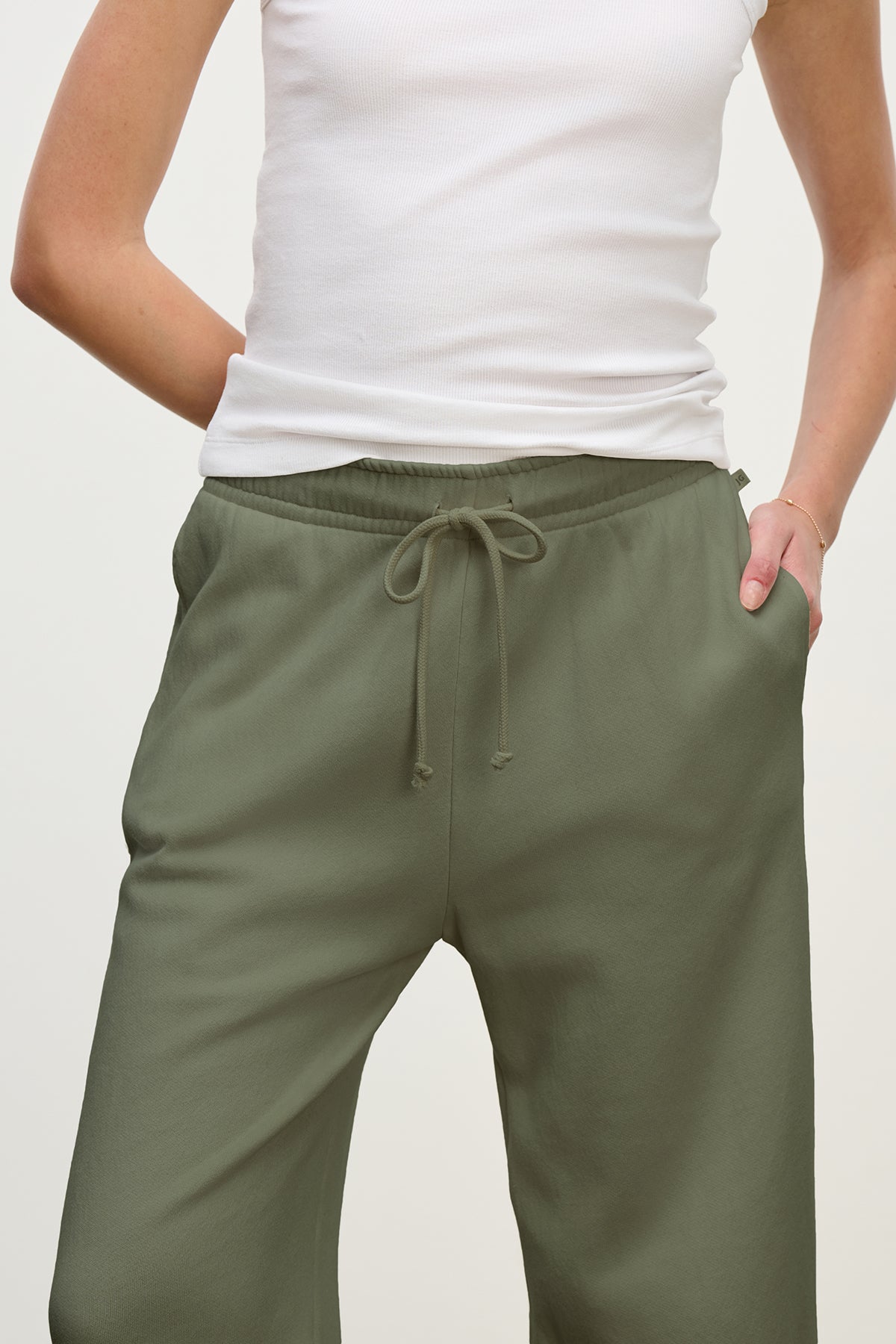   A person wearing MONTECITO SWEATPANT from Velvet by Jenny Graham in olive-green, paired with a white sleeveless top, standing with hands in pockets against a plain background. 