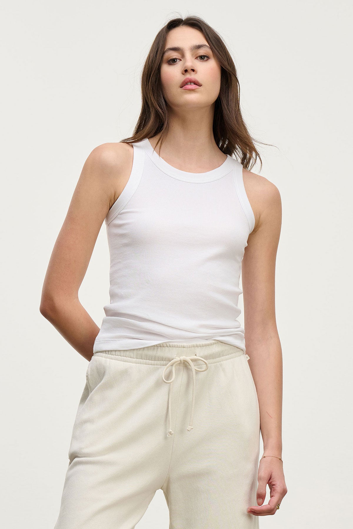 A woman stands against a plain background wearing the CRUZ TANK TOP in white by Velvet by Jenny Graham and light beige drawstring sweatpants—essentials for any capsule wardrobe.-39195412332737