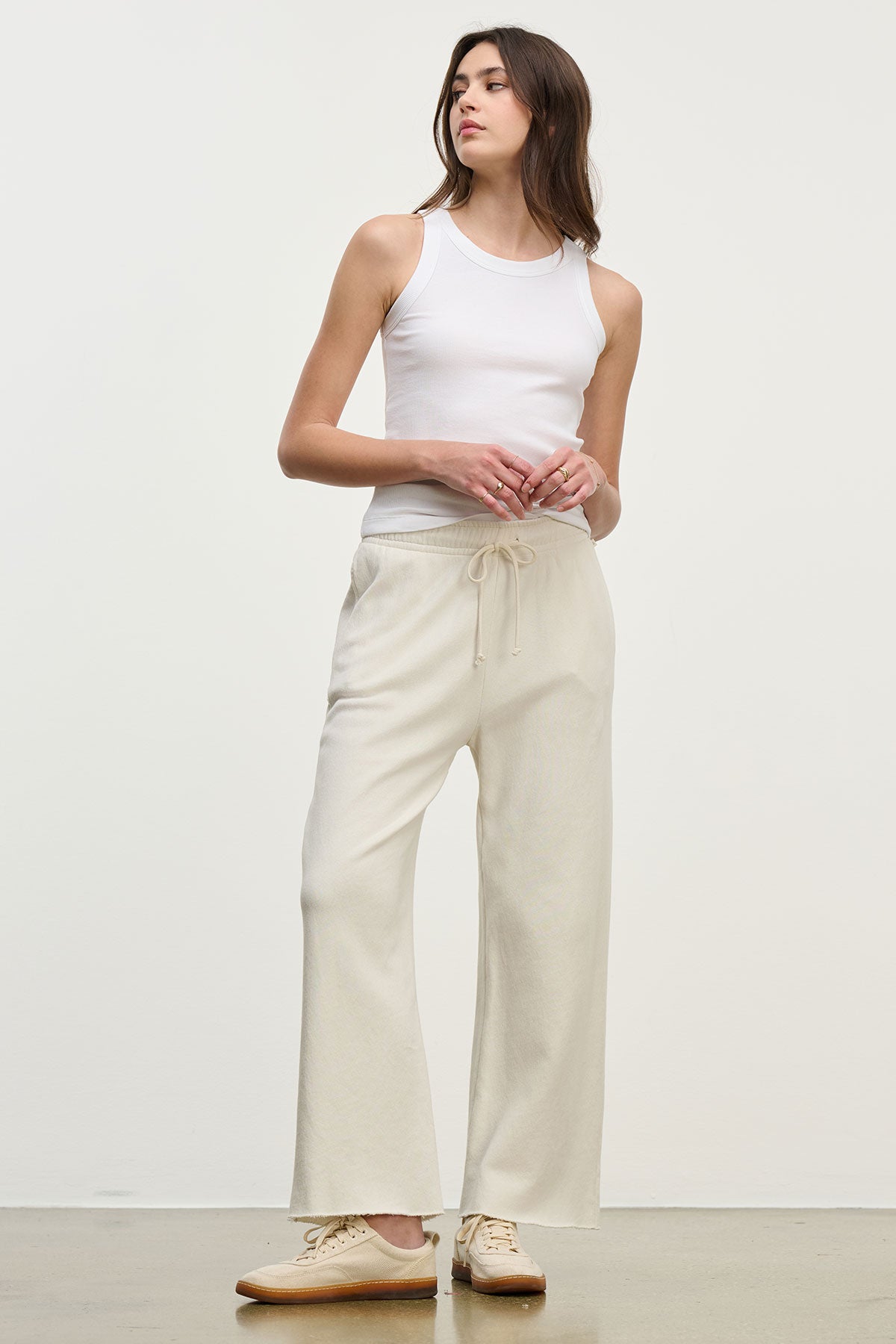   A person in a white tank top and MONTECITO SWEATPANT by Velvet by Jenny Graham, in light beige, stands against a plain background, looking to the side. 