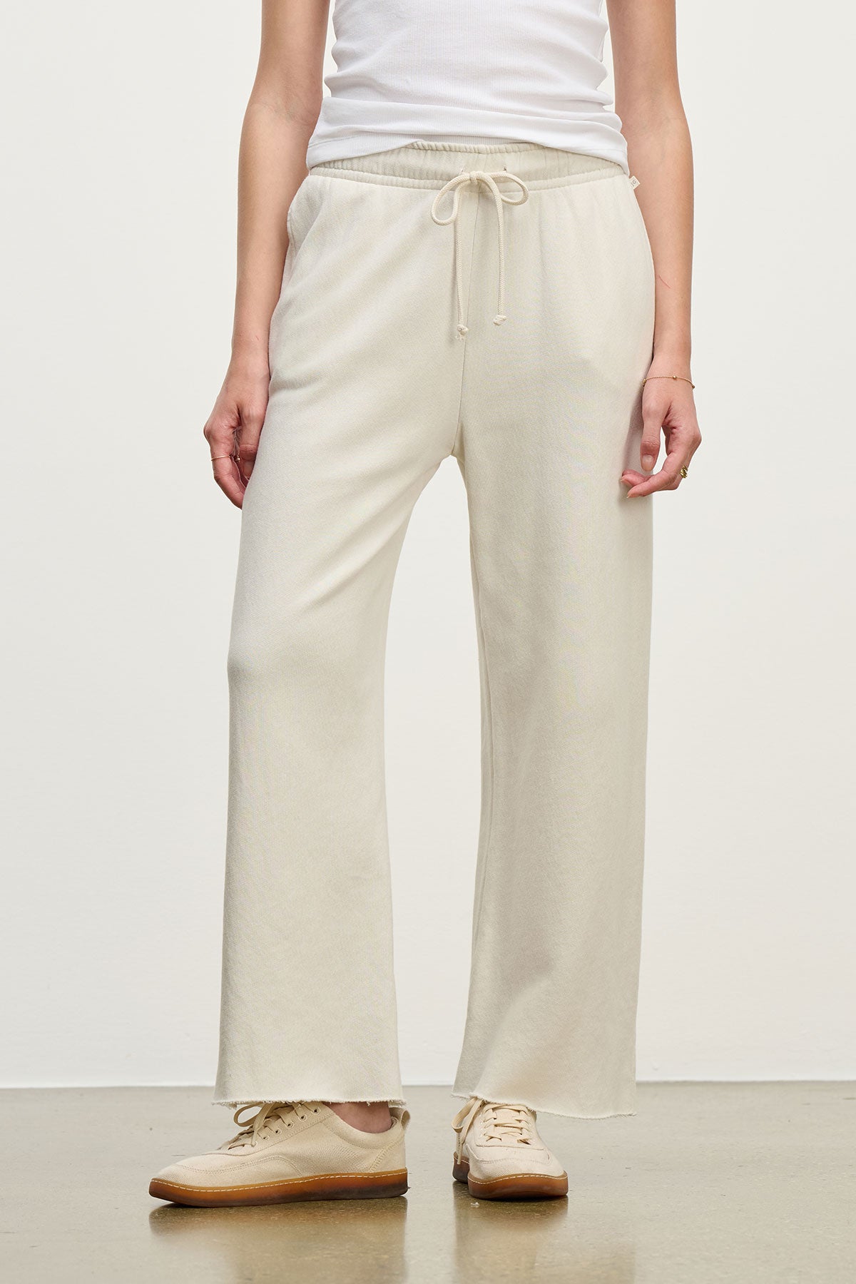   A person in MONTECITO SWEATPANT by Velvet by Jenny Graham, cream-colored relaxed-fit joggers crafted from organic cotton, paired with a white top and beige sneakers against a plain background. 