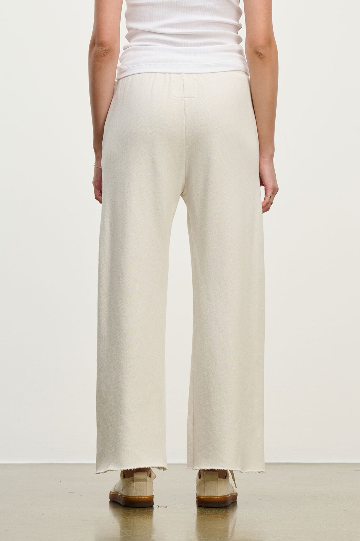   A person wears MONTECITO SWEATPANT by Velvet by Jenny Graham, a cream-colored flared-leg pant made from organic cotton, and a white sleeveless top. They stand with their back to the camera against a plain background. 