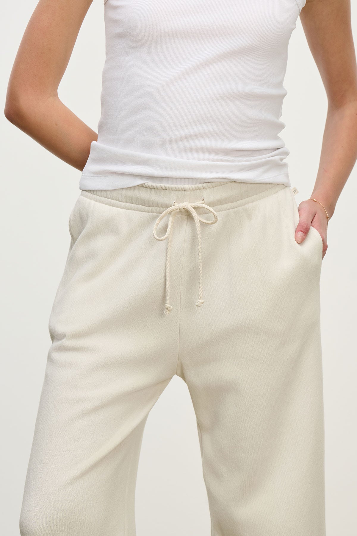   Someone wearing the MONTECITO SWEATPANT by Velvet by Jenny Graham, a white sleeveless top with cream-colored, relaxed-fit joggers made of organic cotton stands against a plain background, one hand in their pocket. 