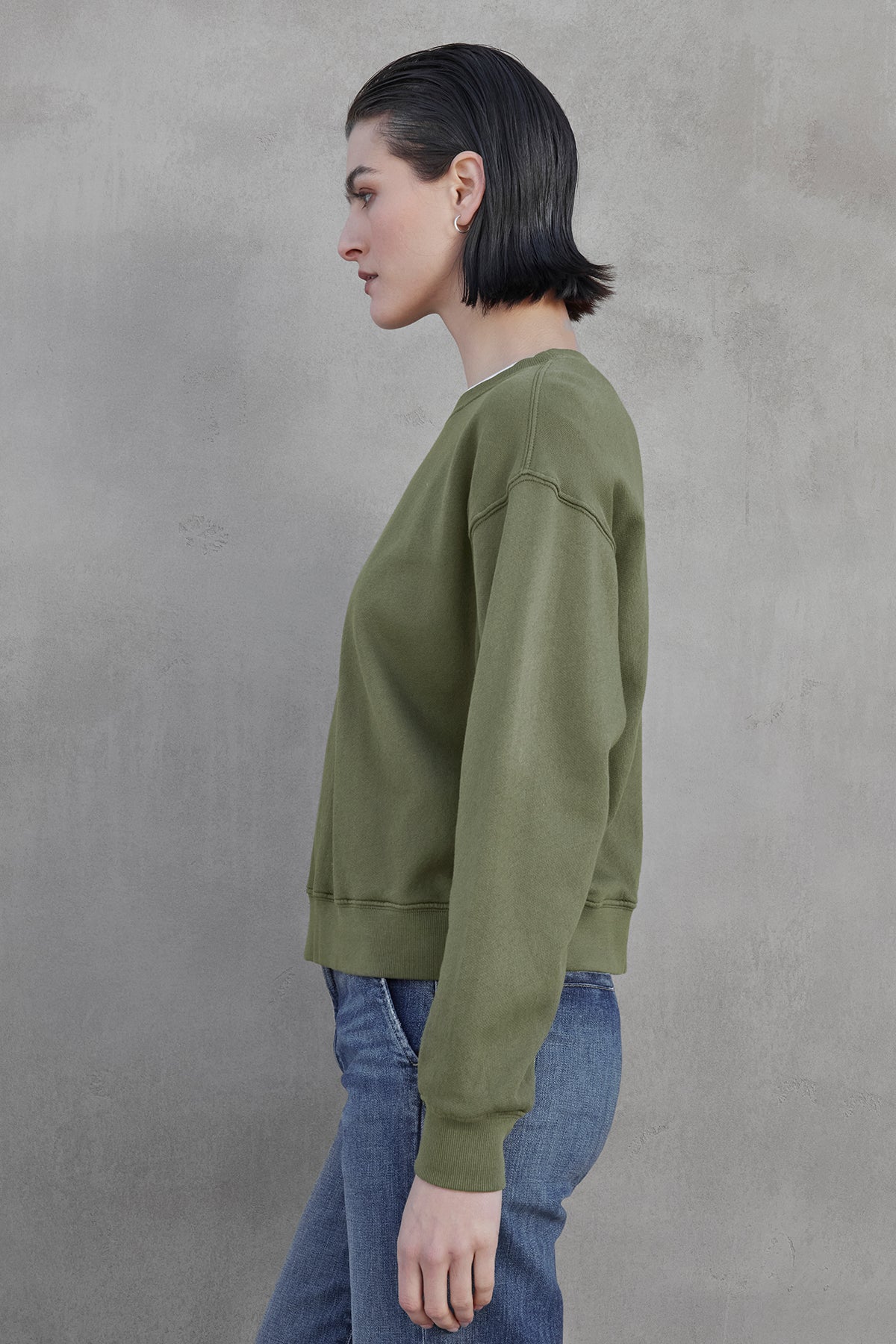   A woman with short dark hair stands sideways against a gray background, wearing Velvet by Jenny Graham’s YNEZ SWEATSHIRT in green organic fleece and blue jeans, embodying sustainable fashion. 