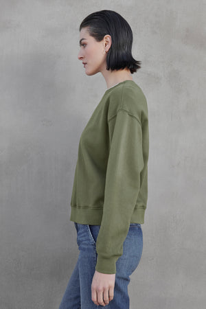 A woman with short dark hair stands sideways against a gray background, wearing Velvet by Jenny Graham’s YNEZ SWEATSHIRT in green organic fleece and blue jeans, embodying sustainable fashion.