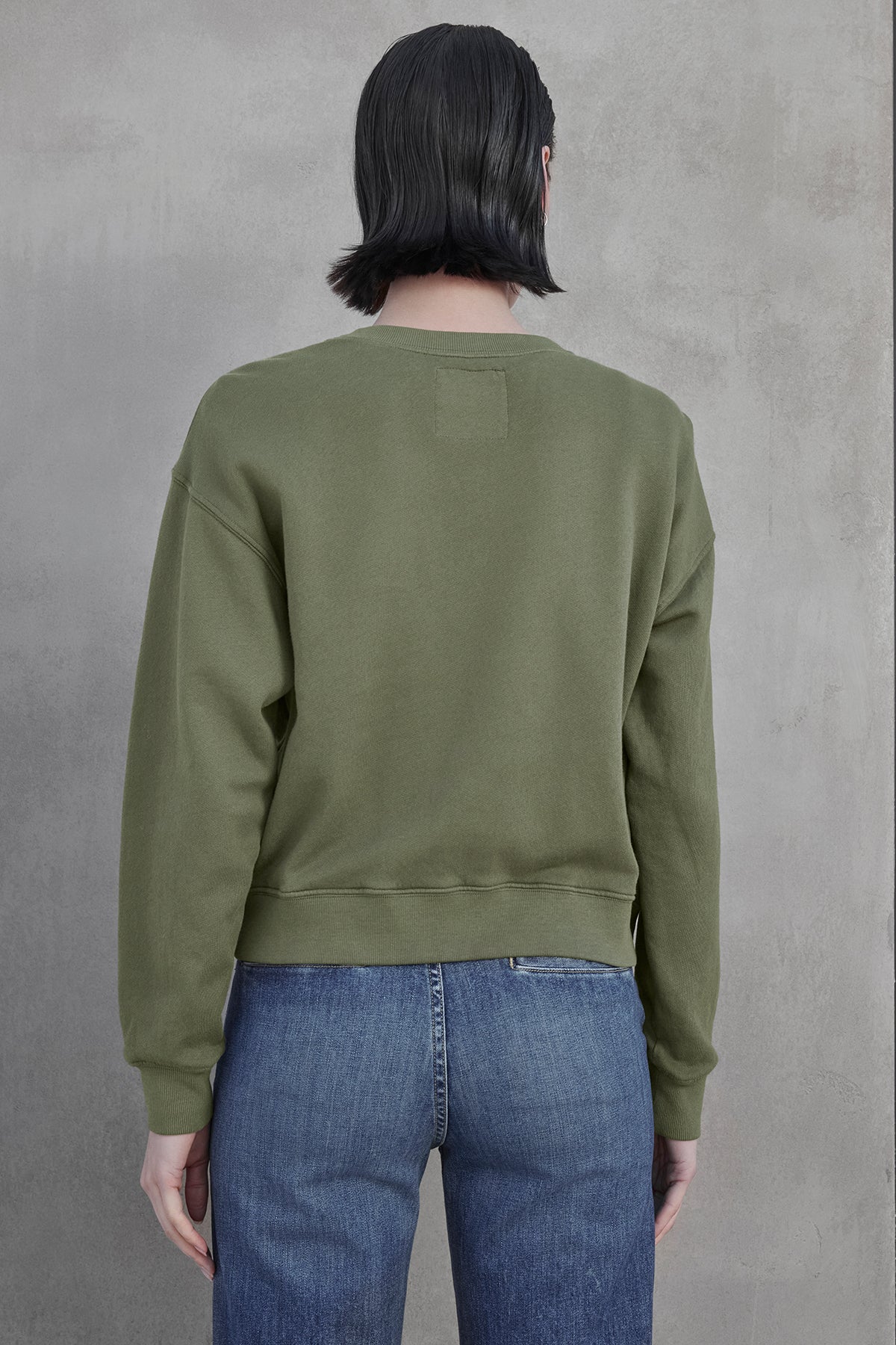   A person with shoulder-length dark hair is wearing a YNEZ SWEATSHIRT by Velvet by Jenny Graham made from organic cotton and blue jeans, standing with their back to the camera. 