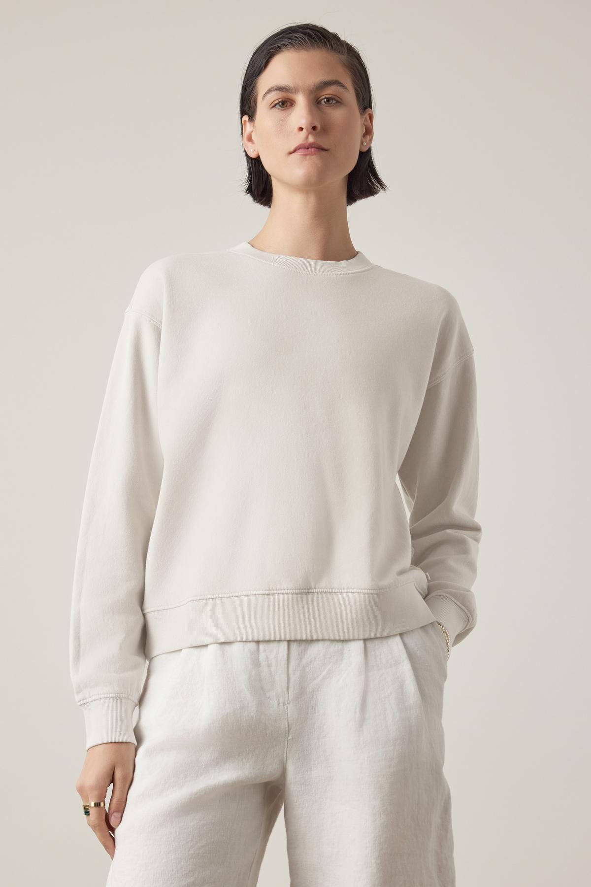   Person wearing a Velvet by Jenny Graham YNEZ SWEATSHIRT made of organic cotton and white pants, standing against a plain background with a neutral expression. 