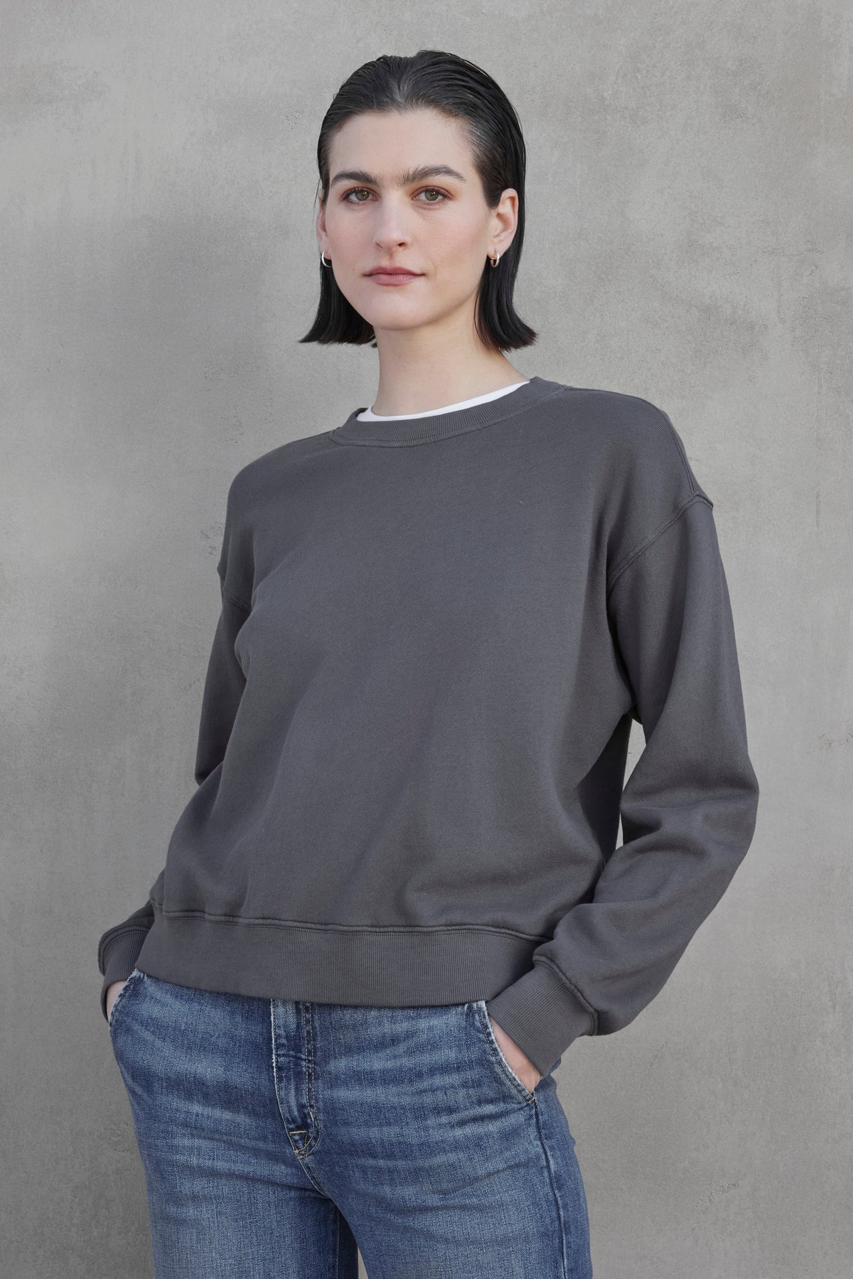   A person with short dark hair, dressed in the YNEZ SWEATSHIRT from Velvet by Jenny Graham and blue jeans, stands with hands in pockets against a plain gray background. 