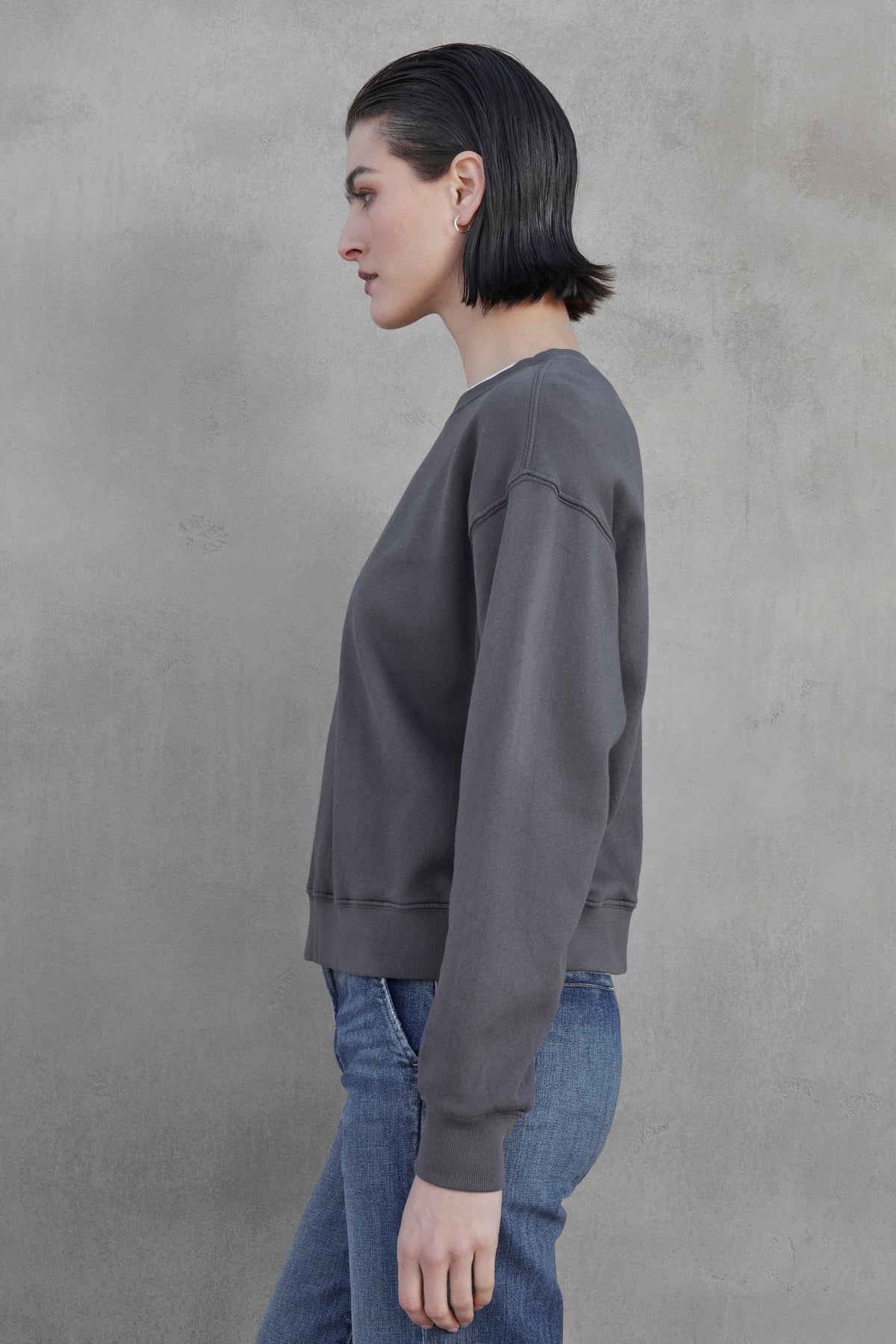   A person with short, dark hair is standing sideways wearing the YNEZ SWEATSHIRT by Velvet by Jenny Graham, a dark gray sweatshirt made of environmentally friendly organic cotton, paired with blue jeans against a plain gray background. 